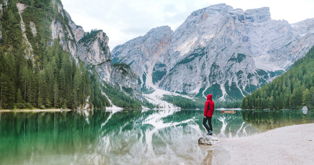 Person standing at a lake with mountains in the background.