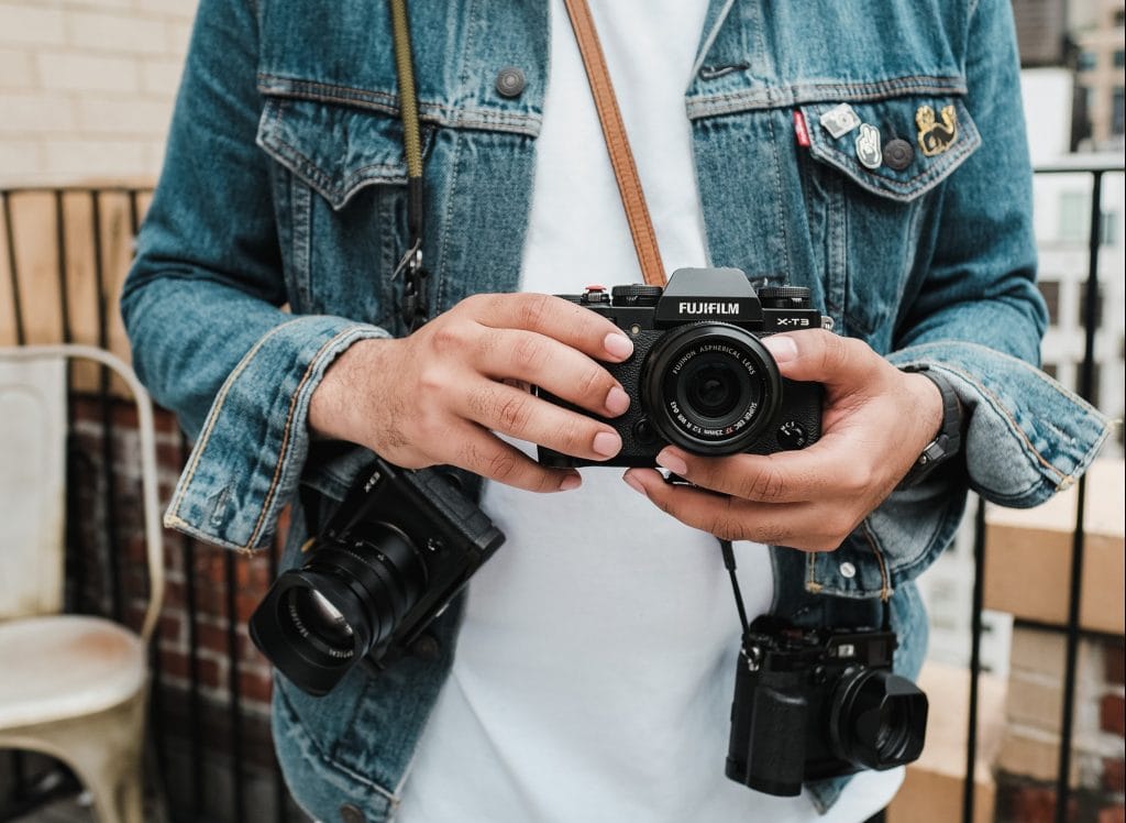 Person in denim jacket wearing multiple cameras around their neck and shoulders, with a Fujifilm X-T3 center and in their hands.
