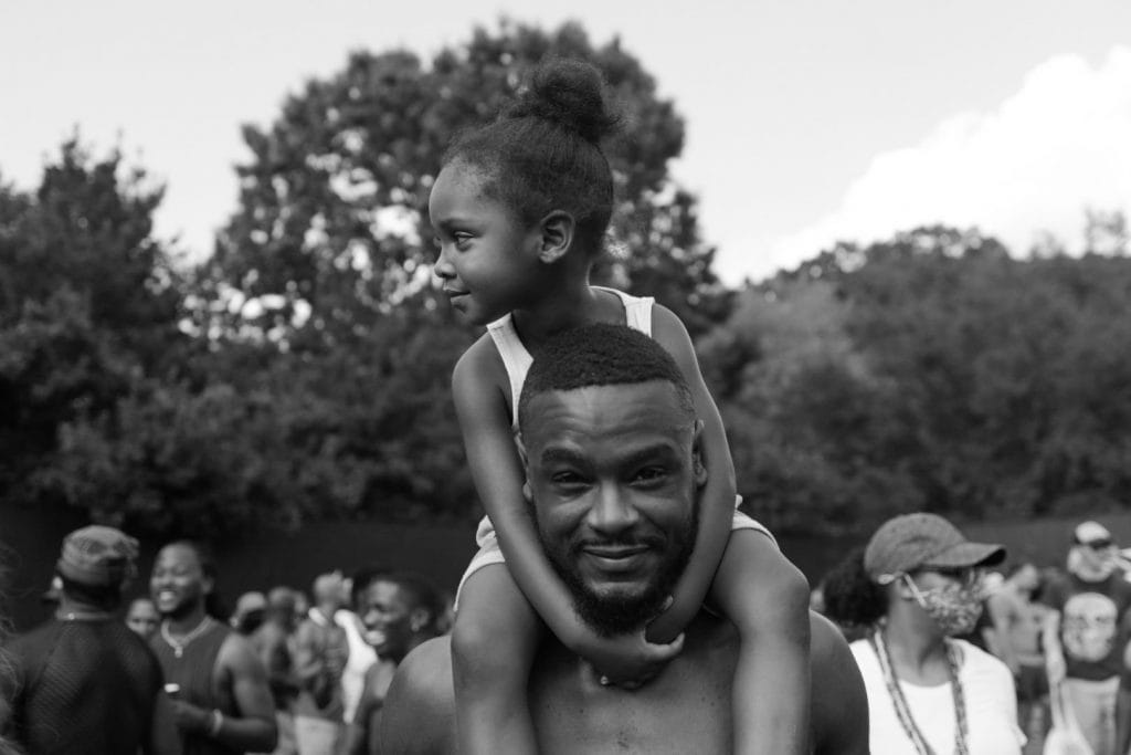 Black and white photo of a young Black girl sitting on her father's shoulders.