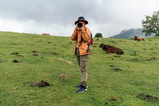 Wildlife photographer pointing camera straight ahead with grassy hill and cows behind them.