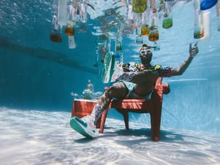 Fashion model wearing sunglasses sitting in chair underwater surrounded by floating bottles