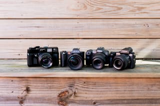 Photo of 4 black cameras from different brands sitting on a wooden bench